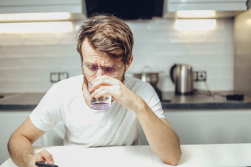 young shaggy man drinks alcohol in the kitchen with a funny face. husband relaxes