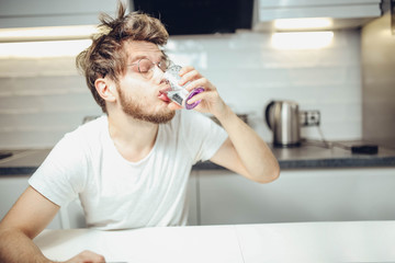 young shaggy man drinks alcohol in the kitchen with a funny face. husband relaxes