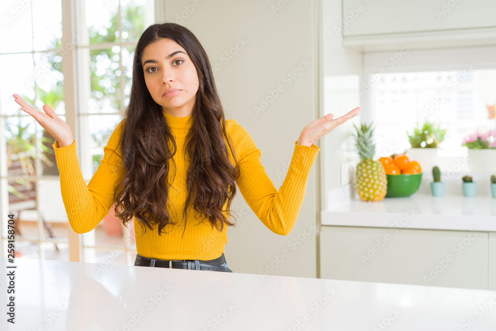 Poster Young beautiful woman at home on white table clueless and confused expression with arms and hands raised. Doubt concept.