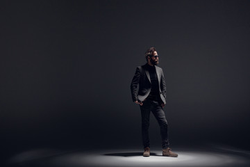 Handsome bearded man wearing in black suit, poses in profile in studio, on a dark lightt background.