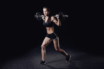Young athletic woman doing barbell squats in gym, black isolated background