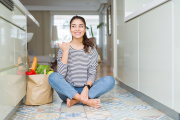 Young woman sitting on the kitchen floor with a paper bag full of fresh groceries smiling with happy face looking and pointing to the side with thumb up.