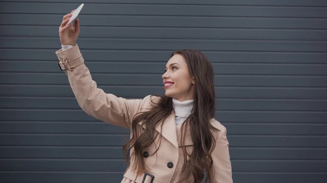 smiling elegant woman in beige trench coat taking selfie on smartphone in windy weather