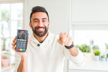 Handsome hispanic man holding point of sale terminal dataphone pointing with finger to the camera...