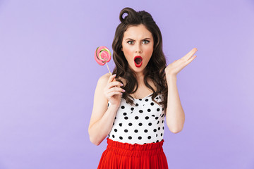 Portrait of happy pin-up woman 20s in vintage polka dot dress smiling while holding and eating colorful lollipop