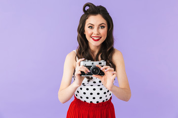 Portrait of brunette pin-up woman 20s in vintage polka dot dress smiling while holding retro photo camera