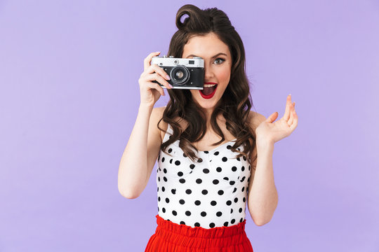 Portrait of seductive pin-up woman 20s in vintage polka dot dress smiling while holding retro photo camera