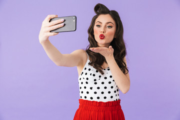 Photo of caucasian pin-up woman 20s in vintage polka dot dress holding and taking selfie photo on black smartphone