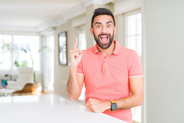 Handsome hispanic man wearing casual t-shirt at home pointing finger up with successful idea. Exited and happy. Number one.