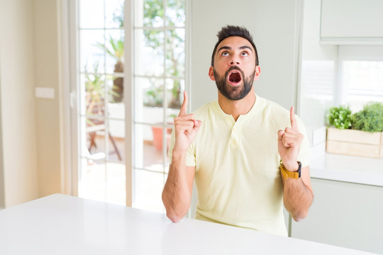 Handsome hispanic man casual yellow t-shirt at home amazed and surprised looking up and pointing with fingers and raised arms.