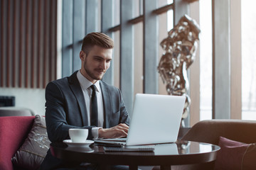 Young handsome man sitting in office with cup of coffee and working on project connected with modern cyber technologies. Businessman with notebook trying to keep deadline in digital marketing sphere.