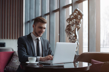 Young handsome man sitting in office with cup of coffee and working on project connected with modern cyber technologies. Businessman with notebook trying to keep deadline in digital marketing sphere.