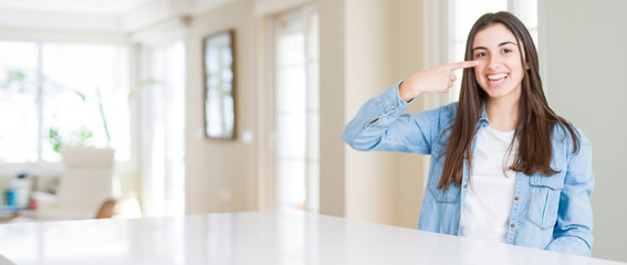 Wide angle picture of beautiful young woman sitting on white table at home Pointing with hand finger to face and nose, smiling cheerful