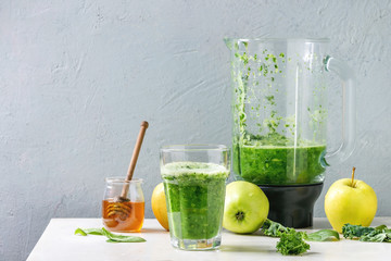 Green spinach kale apple honey smoothie in glass on white marble table with blender and ingredients above. Healthy organic eating.