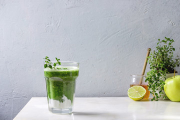 Green spinach kale apple honey smoothie with yogurt and thyme in glass on white marble table with ingredients above. Healthy organic eating.