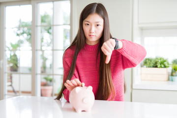 Beautiful Asian woman putting a coin inside piggy bag with angry face, negative sign showing dislike with thumbs down, rejection concept