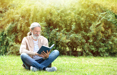 Handsome mature man sitting on green grass and reading book in park, space for text