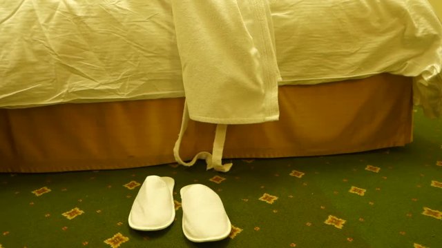 White bathrobe and disposable slippers on unmade bed in hotel room lit by warm golden light
