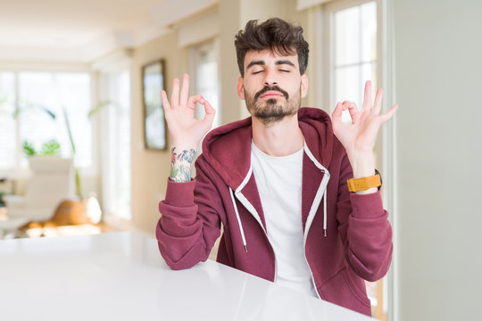 Young man wearing casual sweatshirt sitting on white table relax and smiling with eyes closed doing meditation gesture with fingers. Yoga concept.