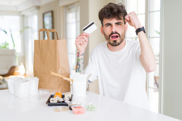 Young man eating asian sushi from home delivery using credit card as payment annoyed and frustrated shouting with anger, crazy and yelling with raised hand, anger concept