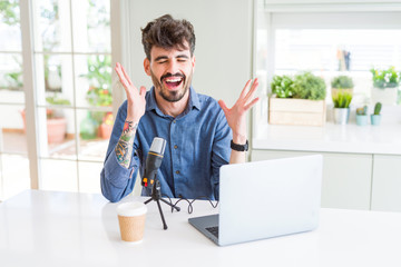 Young man recording podcast using microphone and laptop celebrating mad and crazy for success with arms raised and closed eyes screaming excited. Winner concept