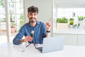 Young man recording podcast using microphone and laptop very happy pointing with hand and finger to the side