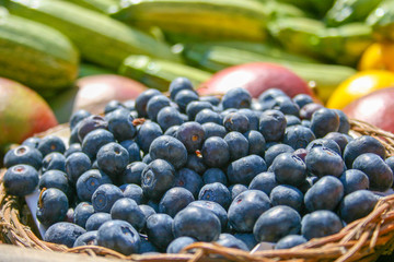 Fresh organic ripe blueberries in a basket with other blurred out of focus fruits and vegetables on the background, in a market, close up
