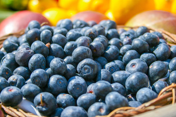Fresh organic ripe blueberries in a basket with other blurred out of focus fruits and vegetables on the background, in a market, close up
