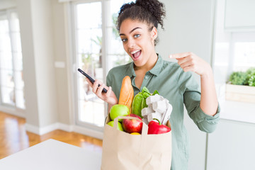 Young african american girl holding paper bag of groceries and using smartphone very happy pointing with hand and finger
