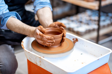 Man making clay jug forming shape by hands on the pottery wheel indoors, close-up view