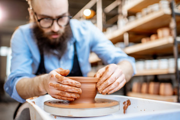 Handsome man as a potter worker in apron making clay jugs on the pottery wheel at the small...