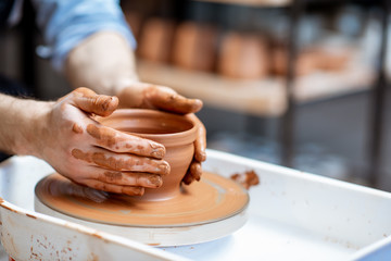 Man making clay jug forming shape by hands on the pottery wheel indoors, close-up view