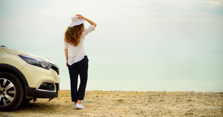 Happy stylish young woman traveler on beach road near white crossover car, holding hat in hand. Banner. Travel, summer vacation, holiday, freedom concept. Digital detox