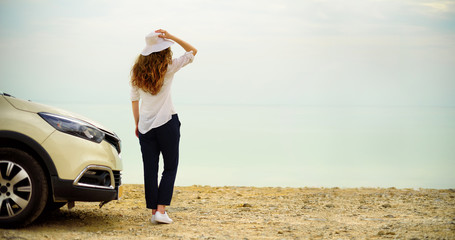 Fototapeta premium Happy stylish young woman traveler on beach road near white crossover car, holding hat in hand. Banner. Travel, summer vacation, holiday, freedom concept. Digital detox
