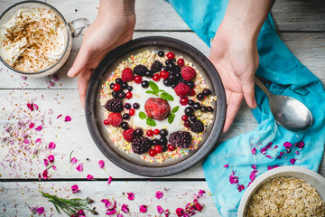 granola, fresh, nutritious breakfast with the addition of natural yoghurt, fresh and frozen fruit, and mint leaves, served on a plate held by female hands, on a white table decorated with spring flowe