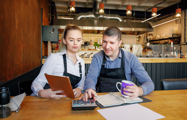 Young woman and her husband sitting at table with tablet and crumpled papers doing accounts for their small restaurant late in evening. Restaurant owners. Start-up entrepreneurs