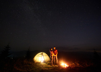 Romantic couple hikers man and woman standing in front of brightly lit tent by burning campfire, holding hands under starry sky, enjoying quiet night camping in mountains. Tourism, traveling concept.