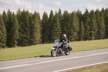 Bearded motorcyclist in helmet, sunglasses and black leather clothing riding cruiser motorbike down empty asphalt road on sunny summer day on background of tall dense spruce trees forest.