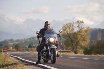 Fototapeta na wymiar Portrait of handsome bearded biker in black leather jacket on cruiser motorcycle on country roadside on blurred background of green woody hills, distant white mountain peaks.