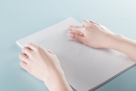 cropped view of young woman reading braille text on paper isolated on blue