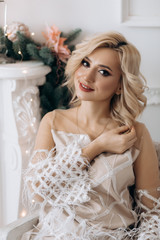 Fototapeta na wymiar Charming blonde woman in white dress poses in a room with large Christmas tree