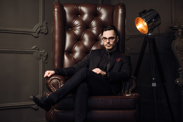 Young handsome man posing for a fashion shoot in a studio. Fashion as a lifestyle. Man wearing a beard. Model sitting on a sofa in tuxedo. Successful fashionable businessman. Business look advertising