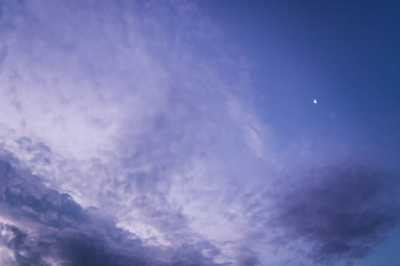 Blue and purple evening sky with different types of clouds like Cirrocumulus, Altocumulus and Cumulus. Small crescent moon behind. 