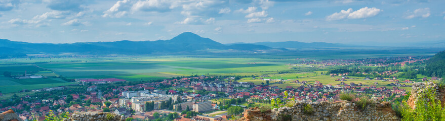 Fototapeta premium Panorama of Rasnov city and small villages in Transylvania, Romania, Eastern Europe. Green fields, traditional houses, hills and cloudy sky photographed in a summer day from a medieval fortress.