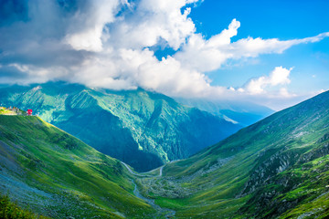 Transfagarasan road in the Carpathian Mountains, Romania, Eastern Europe. Alpine landscape with green meadows, blue sky, dramatic clouds and red cottage. 