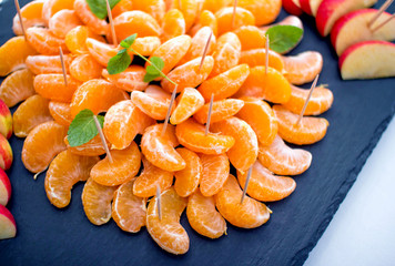 Peeled tangerine slices and apple skewers, served on black stone tray, decorated with mint leaves and sugar powder.