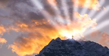 Silhouette of a cross on the top of a mountain with sun rays and dramatic clouds in the background....