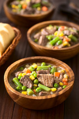 Beef stew or soup with colorful summer vegetables (pea, carrot, sweet corn, green bean, onion), photographed on dark wood with natural light (Selective Focus, Focus in the middle of the first dish)