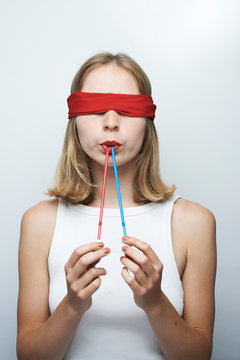 Young blindfold woman holding straw in mouth against gray background