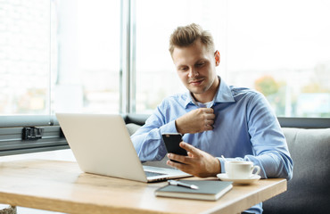 Fototapeta na wymiar Young handsome man sitting in office with cup of coffee and working on project connected with modern cyber technologies. Businessman with notebook trying to keep deadline in digital marketing sphere.